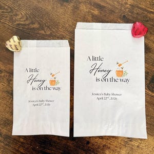 May include: Two white paper favor bags with the text "A little Honey is on the way" and baby shower details. Each bag has a honey pot graphic. A gold and a red heart-shaped candy are on the bags.