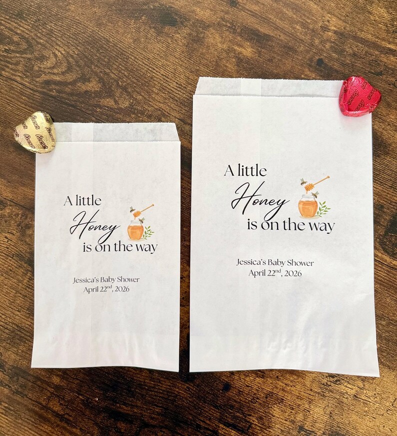 May include: Two white paper favor bags with the text "A little Honey is on the way" and baby shower details. Each bag has a honey pot graphic. A gold and a red heart-shaped candy are on the bags.