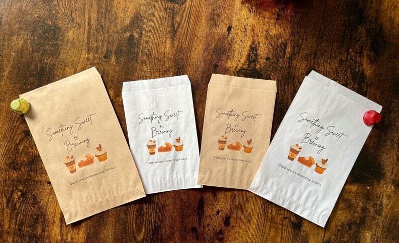 May include: Four paper bags with the text "Something Sweet is Brewing" and illustrations of coffee cups, bread, and cupcakes. The bags are in shades of brown and white, with a wooden background.