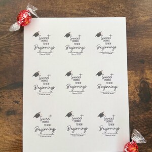 K&ouml;nnte beinhalten: Ein Bogen wei&szlig;er Aufkleber mit dem Text "Sweet Endings to New Beginnings" und einer Abschlusskappe. Die Aufkleber sind f&uuml;r den Jahrgang 2020. Zwei rot-silberne Bonbons liegen auf der Holzoberfl&auml;che.