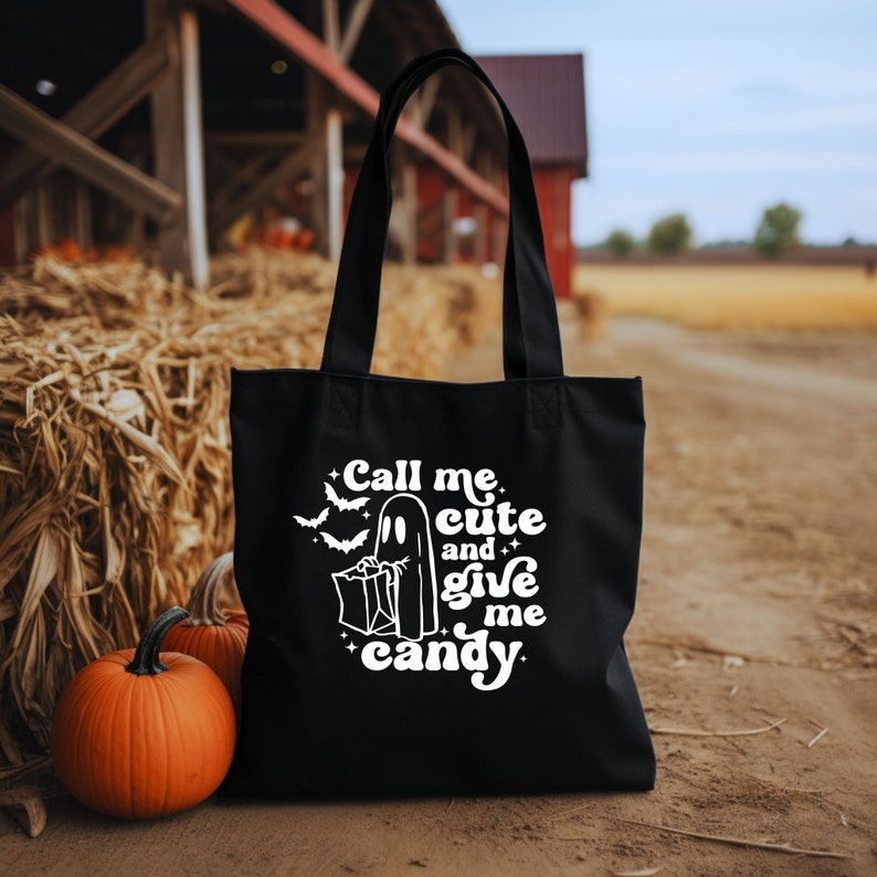 May include: Black tote bag with white text reading "Call me cute and give me candy", featuring a ghost and bats graphic. Two orange pumpkins are in front of the bag. The background includes a hay bale and a red barn.