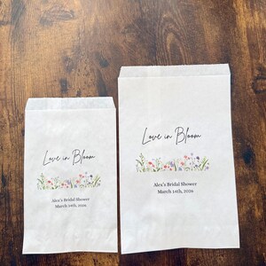 May include: Two white paper bags with the text "Love in Bloom" in elegant script. Below the text is a floral design with red, purple, and green accents. The bags also include the text "Alex's Bridal Shower March 14th, 2026."