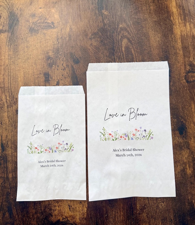 May include: Two white paper bags with the text "Love in Bloom" in elegant script. Below the text is a floral design with red, purple, and green accents. The bags also include the text "Alex's Bridal Shower March 14th, 2026."