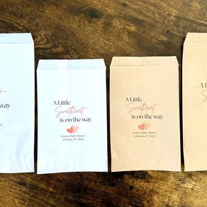 May include: Four paper favour bags of different sizes, arranged on a wooden surface. The bags are white and tan, each printed with the text "A Little Sweetheart is on the way" in a script font, with two pink hearts.