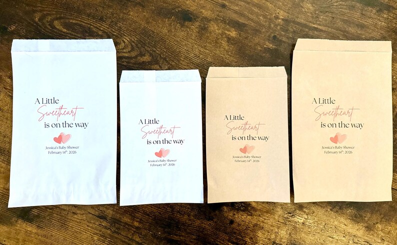 May include: Four paper favour bags of different sizes, arranged on a wooden surface. The bags are white and tan, each printed with the text "A Little Sweetheart is on the way" in a script font, with two pink hearts.