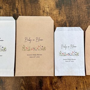 以下が含まれることがあります： 様々なサイズの4つの紙製のギフトバッグ。テキスト「Baby in Bloom」と花のイラストが描かれています。バッグは白と茶色で、「Jessica's Baby Shower June 28, 2025」のテキストが入っています。