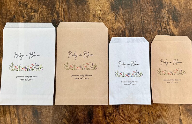 以下が含まれることがあります： 様々なサイズの4つの紙製のギフトバッグ。テキスト「Baby in Bloom」と花のイラストが描かれています。バッグは白と茶色で、「Jessica's Baby Shower June 28, 2025」のテキストが入っています。