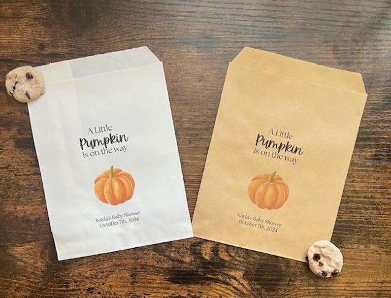 May include: Two paper treat bags, one white and one brown, with a pumpkin theme. Each bag has the text "A Little Pumpkin is on the way" and a watercolor pumpkin illustration. The white bag has a chocolate chip cookie next to it. The brown bag has a chocolate chip cookie next to it. The text "Kayla's Baby Shower October 7th 2024" is printed on the bags.