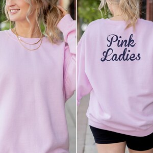 May include: A light pink sweatshirt with the text "Pink Ladies" in navy blue on the back. The sweatshirt is a crew neck style and appears to be made of a soft material. The front of the sweatshirt is plain.