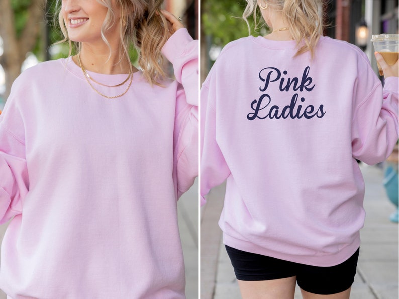 May include: A light pink sweatshirt with the text "Pink Ladies" in navy blue on the back. The sweatshirt is a crew neck style and appears to be made of a soft material. The front of the sweatshirt is plain.