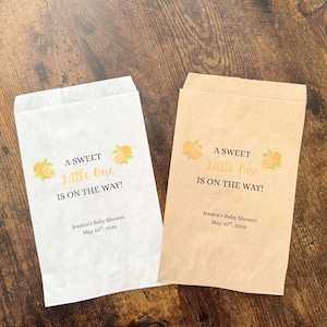 K&ouml;nnte beinhalten: Zwei Papiert&uuml;ten, eine wei&szlig;e und eine beige, mit dem Text "A SWEET Little One IS ON THE WAY!" und Orangenillustrationen. Die T&uuml;ten sind f&uuml;r Jessicas Babyparty, 10. Mai 2026.