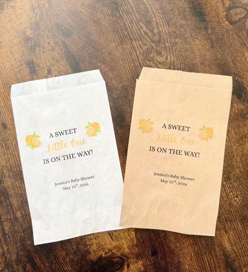 K&ouml;nnte beinhalten: Zwei Papiert&uuml;ten, eine wei&szlig;e und eine beige, mit dem Text "A SWEET Little One IS ON THE WAY!" und Orangenillustrationen. Die T&uuml;ten sind f&uuml;r Jessicas Babyparty, 10. Mai 2026.