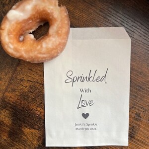 May include: A glazed ring donut and a white paper bag. The donut is light brown with a white glaze. The bag has the words "Sprinkled With Love" and a heart symbol printed on it. The text "Jessica's Sprinkle March 9th 2024" is also printed on the bag.