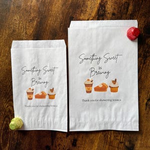 May include: Two white paper treat bags with the text "Something Sweet is Brewing" and illustrations of coffee, pie, and cupcakes. The text "Thank you for showering Jessica" is below the illustrations.