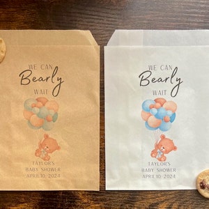 May include: Two paper treat bags, one brown and one white, with a baby shower design. Each bag features a teddy bear holding balloons and the text "We Can Bearly Wait." The date "April 10, 2024" is also printed on the bags.