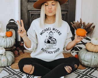 May include: Cream-colored sweatshirt with the text "MICHAEL MYERS BUTCHER SHOP" and a graphic of Michael Myers. The sweatshirt is worn with black ripped jeans. Pumpkins and fall decor are in the background.