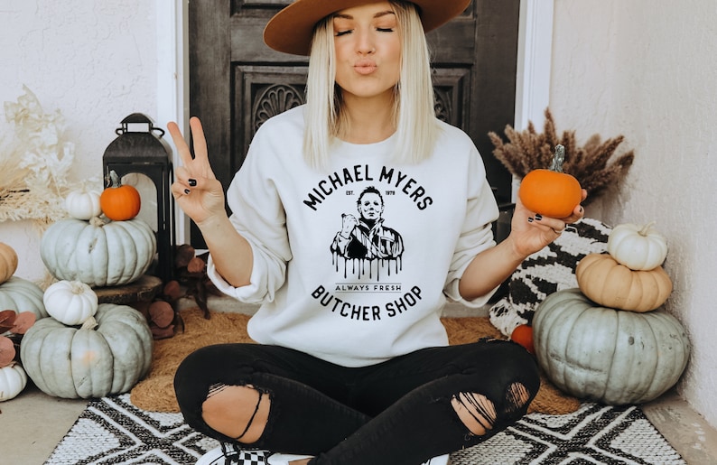 May include: Cream-colored sweatshirt with the text "MICHAEL MYERS BUTCHER SHOP" and a graphic of Michael Myers. The sweatshirt is worn with black ripped jeans. Pumpkins and fall decor are in the background.