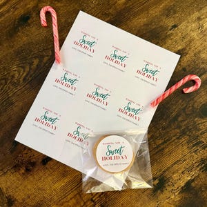 May include: A holiday-themed image featuring a sheet of paper with the text "Wishing You a Sweet Holiday" and a cookie in a clear bag. Two red and white candy canes are placed on the paper. The items are on a wooden surface.