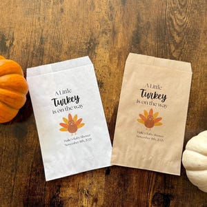 May include: Two paper bags with the text "A Little Turkey is on the way" and a turkey graphic. An orange pumpkin and a white pumpkin are on a wooden surface. Baby shower theme.