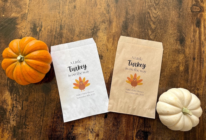 May include: Two paper bags with the text "A Little Turkey is on the way" and a turkey graphic. An orange pumpkin and a white pumpkin are on a wooden surface. Baby shower theme.