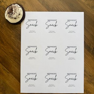 May include: A white rectangular sheet featuring nine identical printed labels, each displaying the text MIDNIGHT Snack in a combination of serif and script fonts, followed by John & Jessica September 8th, 2024. A dark cookie with white frosting sits nearby.