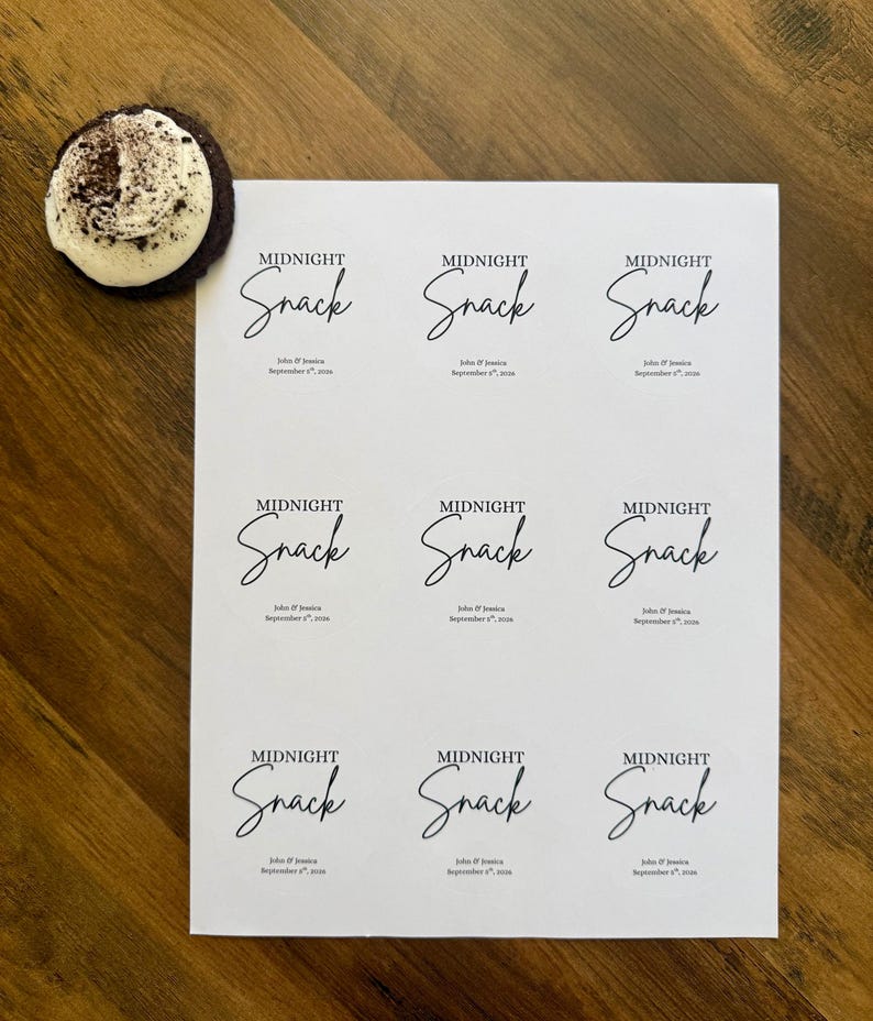 May include: A white rectangular sheet featuring nine identical printed labels, each displaying the text MIDNIGHT Snack in a combination of serif and script fonts, followed by John & Jessica September 8th, 2024. A dark cookie with white frosting sits nearby.