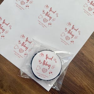 Op de afbeelding: Een vel met ronde witte stickers met rode inkt met de tekst Special Delivery, Jessica's Baby Shower, July 19th, 2026, en iconen van een babyfles, rammelaar, rompertje, hart en twee glazen. E&eacute;n sticker zit in een doorzichtig plastic zakje.