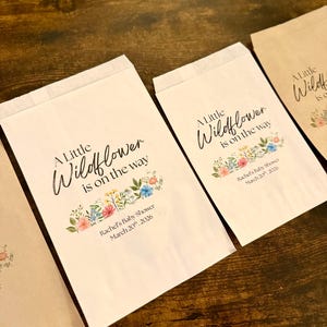 May include: Several paper bags with the text "A Little Wildflower is on the way" and "Rachel's Baby Shower March 20th, 2026". The bags have a floral design in pink, blue, and yellow.