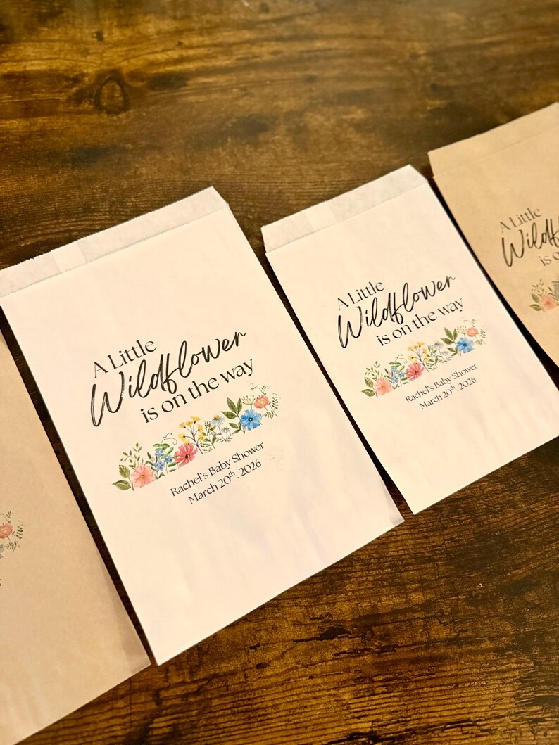 May include: Several paper bags with the text "A Little Wildflower is on the way" and "Rachel's Baby Shower March 20th, 2026". The bags have a floral design in pink, blue, and yellow.