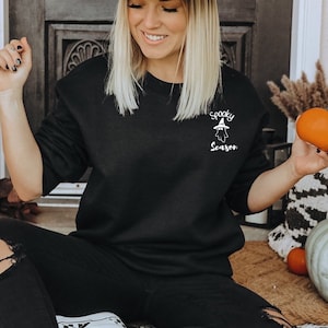 May include: Black sweatshirt with the text "Spooky Season" and a ghost graphic. The sweatshirt is a solid black color and has long sleeves. The text and graphic are in white.