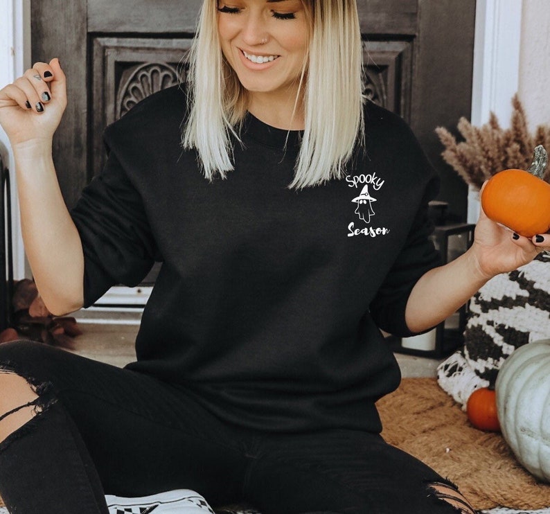 May include: Black sweatshirt with the text "Spooky Season" and a ghost graphic. The sweatshirt is a solid black color and has long sleeves. The text and graphic are in white.