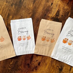 May include: Four paper favor bags with the text "A Baby is Brewing" and a watercolor illustration of coffee cups. The bags are in shades of brown and white, with a baby shower date printed on them.