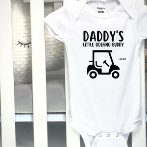 May include: White baby onesie with the text "DADDY'S LITTLE GOLFING BUDDY" in black. A black golf cart graphic is below the text. The onesie is made of soft material and is designed for infants.