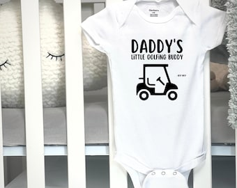 May include: White baby onesie with the text "DADDY'S LITTLE GOLFING BUDDY" in black. A black golf cart graphic is below the text. The onesie is made of soft material and is designed for infants.