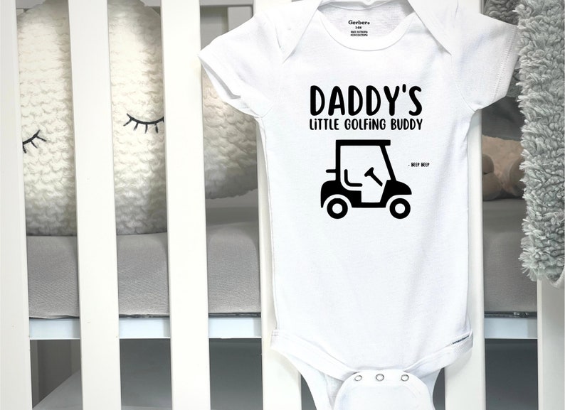 May include: White baby onesie with the text "DADDY'S LITTLE GOLFING BUDDY" in black. A black golf cart graphic is below the text. The onesie is made of soft material and is designed for infants.
