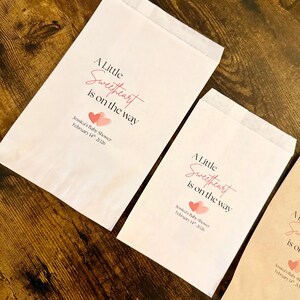 May include: A selection of paper treat bags, each printed with the phrase "A Little Sweetheart is on the way" in pink script. The bags also feature the text "Jessica's Baby Shower February 14th, 2026" and two pink hearts. They rest on a wooden surface.