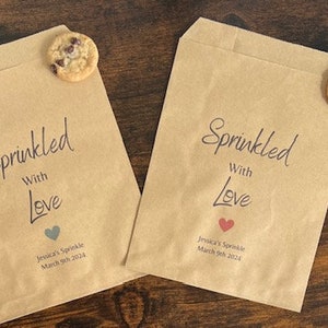 May include: Two brown paper treat bags with the words "Sprinkled With Love" in black script. Each bag features a heart graphic and event details. A chocolate chip biscuit sits on top of each bag.