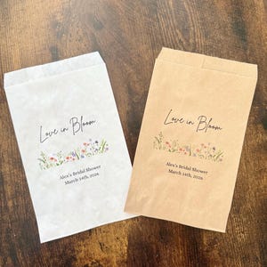 May include: Two paper favor bags, one white and one brown, with the text "Love in Bloom" in cursive. Below the text is a floral design and the text "Alex's Bridal Shower March 14th, 2026."