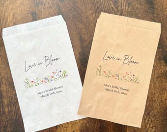 May include: Two paper favor bags, one white and one brown, with the text "Love in Bloom" in cursive. Below the text is a floral design and the text "Alex's Bridal Shower March 14th, 2026."