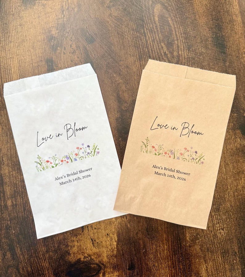 May include: Two paper favor bags, one white and one brown, with the text "Love in Bloom" in cursive. Below the text is a floral design and the text "Alex's Bridal Shower March 14th, 2026."