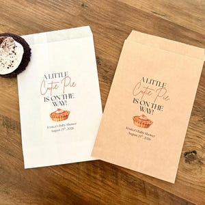May include: Two rectangular paper favor bags, one white and one brown, each printed with the text A LITTLE Cutie Pie IS ON THE WAY! and Jessica's Baby Shower August 25th, 2026, featuring a small pie illustration and a dark cookie with white cream.