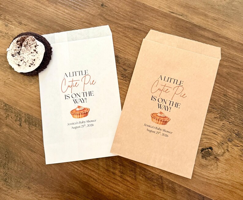 May include: Two rectangular paper favor bags, one white and one brown, each printed with the text A LITTLE Cutie Pie IS ON THE WAY! and Jessica's Baby Shower August 25th, 2026, featuring a small pie illustration and a dark cookie with white cream.