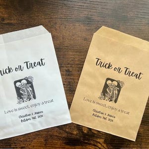 May include: Two Halloween-themed treat bags, one white and one brown, with the text "Trick or Treat". Each bag features a skeleton couple graphic and the message "Love is sweet, enjoy a treat". A cookie rests on each bag.