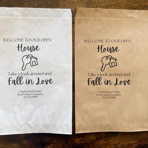 May include: Two paper bags, one white and one tan, with the text "WELCOME TO OUR OPEN HOUSE" and "Take a look around and Fall in Love." A key and house graphic is in the center. The bags also display a real estate company's name and phone number.
