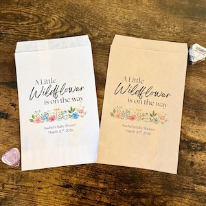 May include: Two paper favor bags, one white and one tan, with the text "A Little Wildflower is on the way." Floral illustrations and event details are printed below the text. Hershey's Kisses are visible.