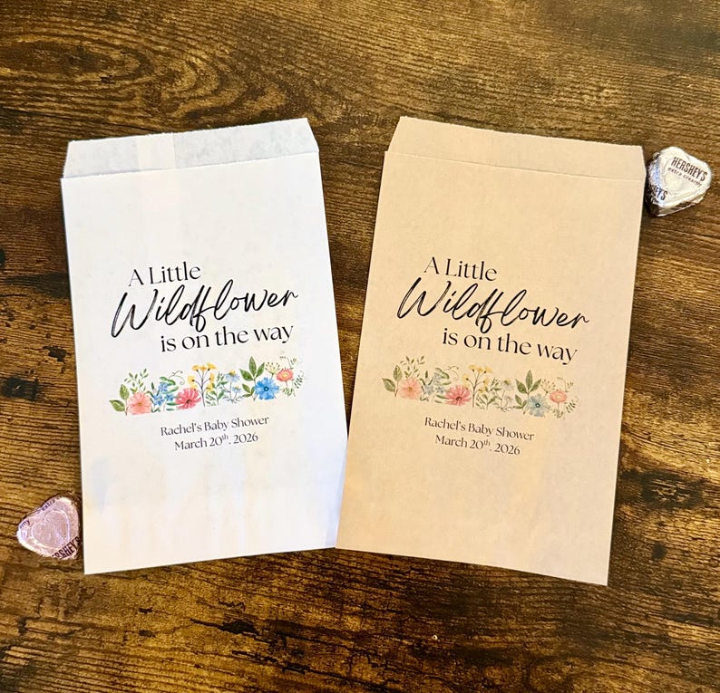 May include: Two paper favor bags, one white and one tan, with the text "A Little Wildflower is on the way." Floral illustrations and event details are printed below the text. Hershey's Kisses are visible.