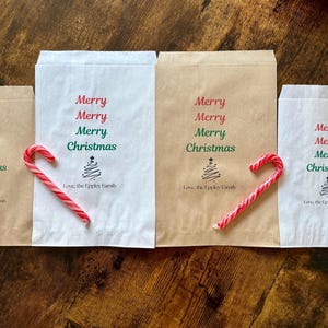 May include: Four paper gift bags, two white and two brown, with "Merry Christmas" printed in red and green. Each bag has a candy cane or a red ornament. The bags are arranged on a wooden surface.