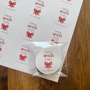 May include: A sheet of circular white stickers featuring the text baking spirits bright in green and red script, accompanied by a red bow illustration and the text Love, The Smith Family. One sticker is individually wrapped in clear plastic.