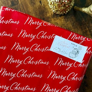 May include: A Christmas gift wrapped in red paper with a repeating white script of "Merry Christmas". A small label reads "Merry Christmas! from Santa Claus" with a Santa illustration. Festive holiday theme.