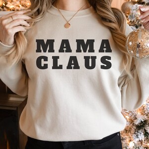 Op de afbeelding: Een cr&egrave;mekleurige sweatshirt met de tekst "MAMA CLAUS" in vette, zwarte letters. De sweatshirt heeft een neutrale kleur en de tekst is gecentreerd. De afbeelding toont een persoon die de sweatshirt draagt.